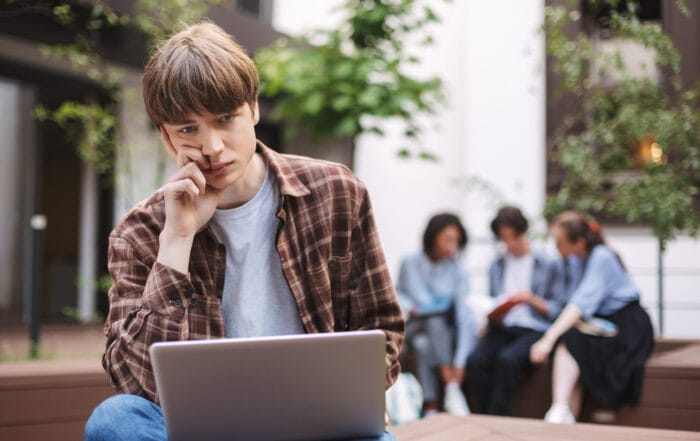 Young boy looking at a laptop, looking lonely and sad. There are a 3 people in the background looking at a phone.
