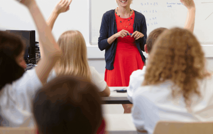 Students in a classroom raising their hands in front of a smiling teacher